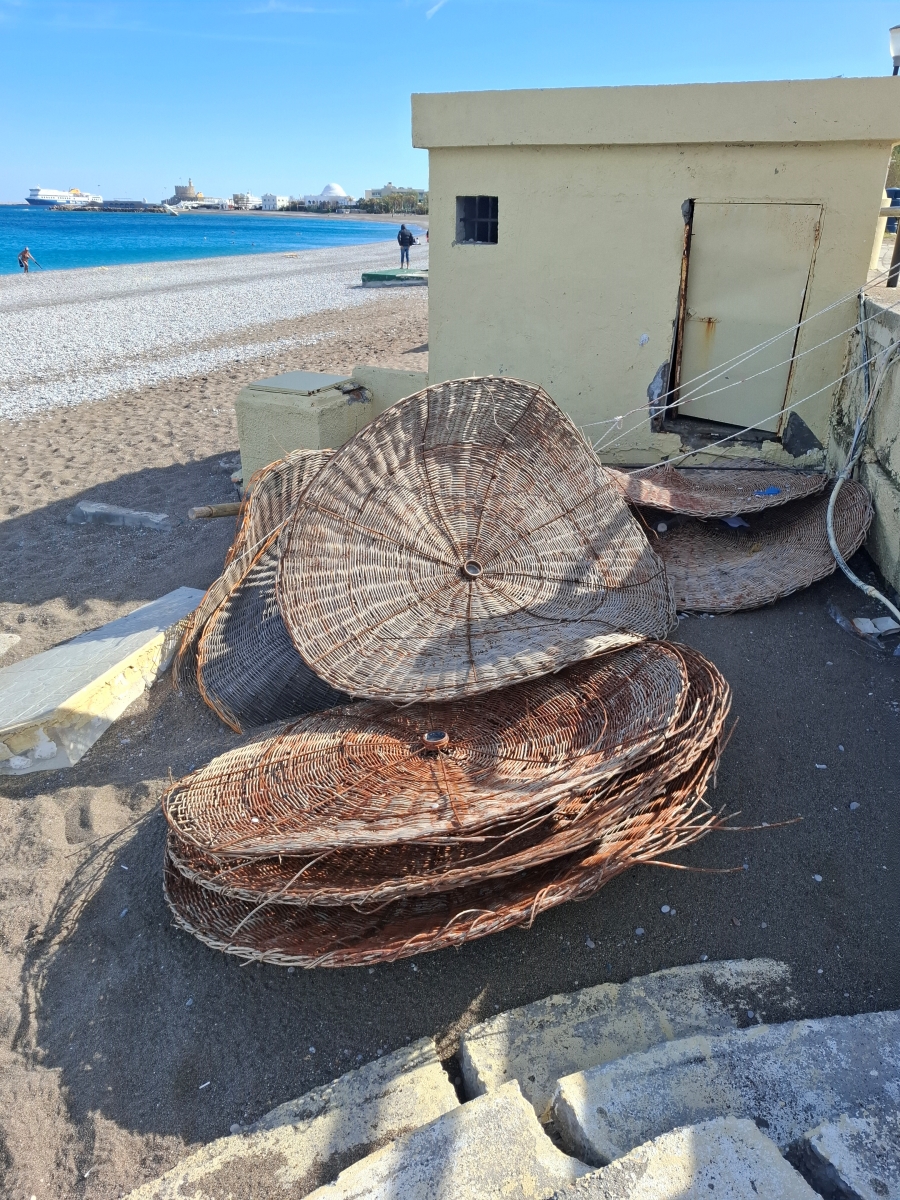 Plecione z wikliny, okrągłe dachy parasoli plażowych leżące na piasku obok jasnego budynku przy plaży.