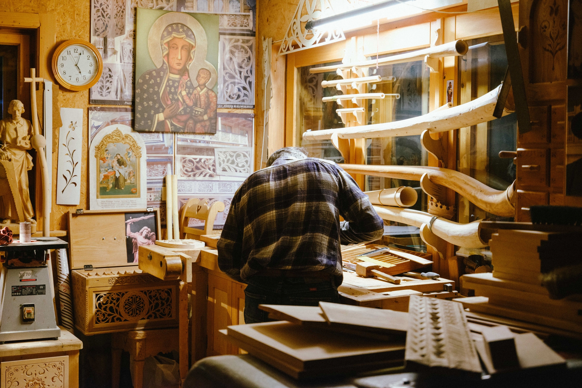 A craftsman, seen from behind, stands over a workbench in his workshop. Around the workshop, there are pieces of wood, works in progress, and finished carvings.