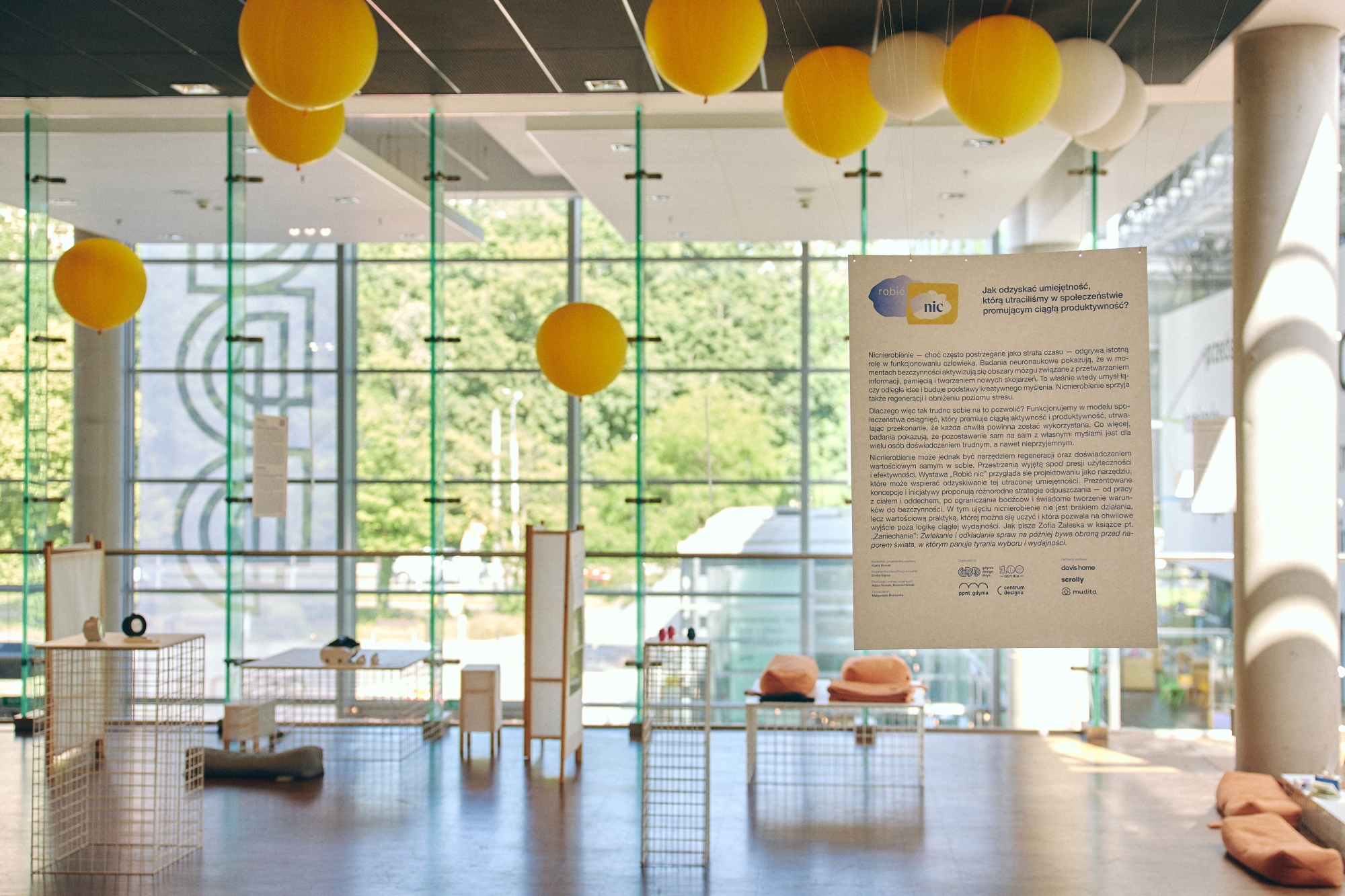A general view of the exhibition space in a bright, glass-walled hall. A hanging board with the exhibition's concept, yellow balloons on the ceiling, and relaxation zones with cushions are visible.