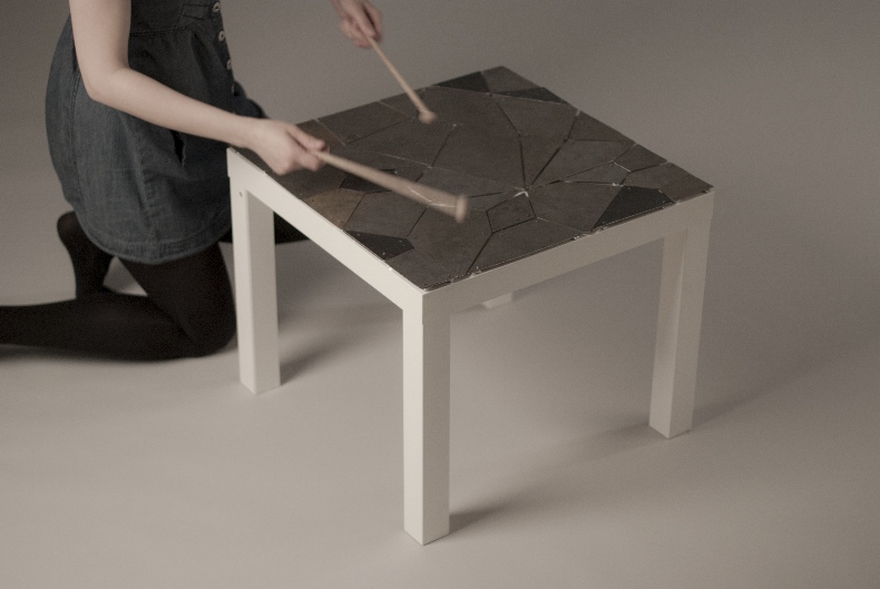 A woman kneels and bangs her chopsticks on the glass top of a wooden coffee table