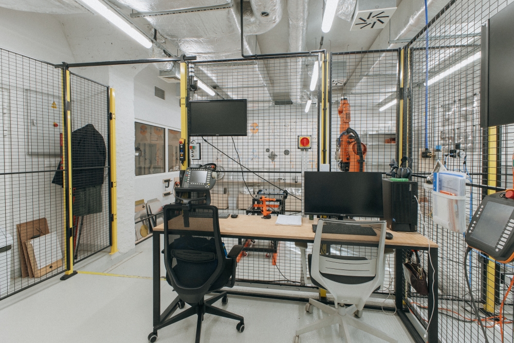 Computer workstation in the KUKA workshop, with chairs and a desk holding a computer in the foreground and a KUKA robotic arm behind a safety fence in the background.