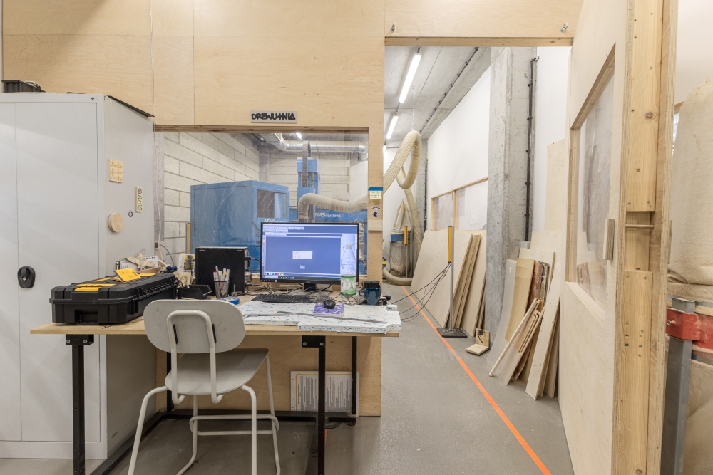 Wnętrze warsztatu stolarskiego. Widok na stanowisko komputerowe, za którym znajduje się kolejne pomieszczenie.