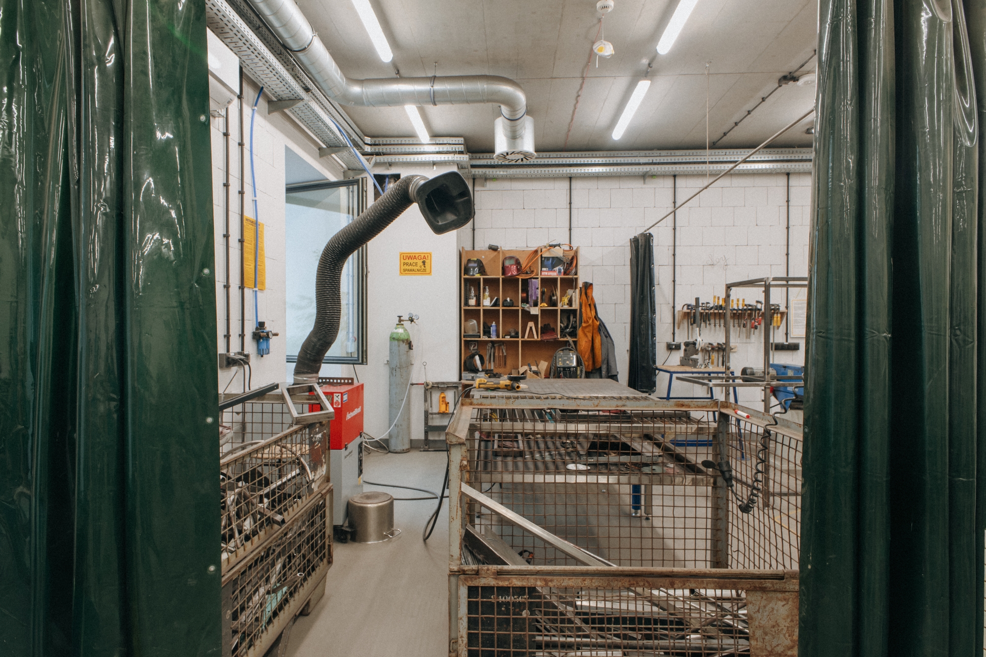 Wnętrze warsztatu ślusarskiego. Za rozsuniętą kotarą widoczne rury i metalowe przedmioty.