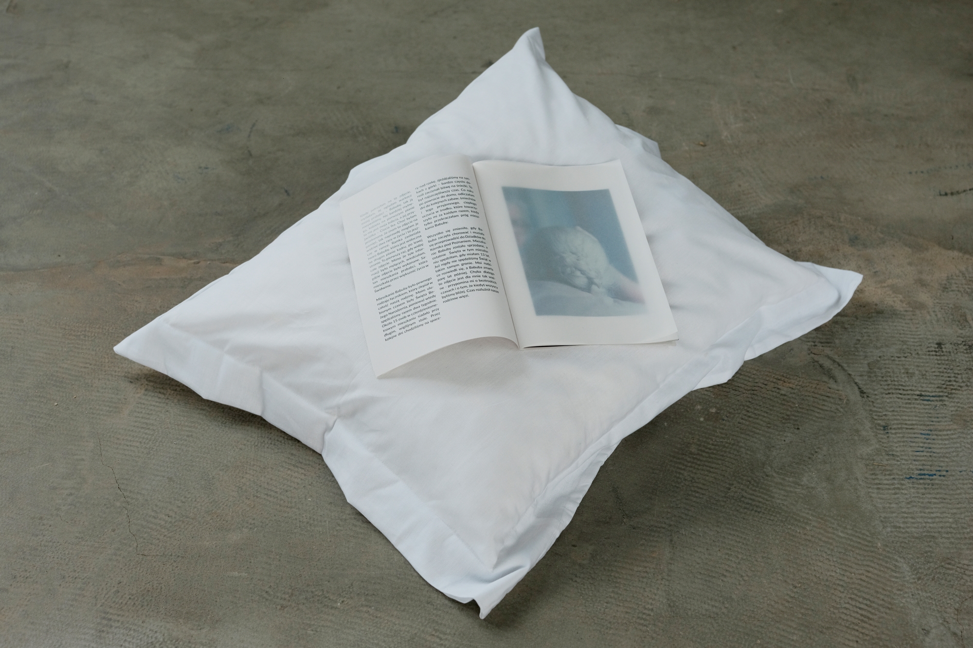 A photographic publication designed by Maria Sadowska is positioned on a pillow on a concrete floor.