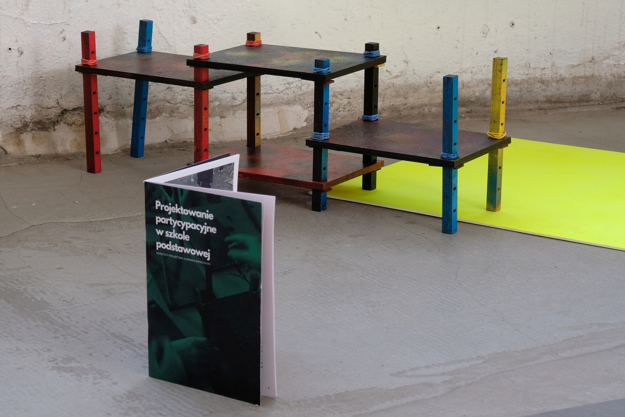 Na pierwszym planie papierowa publikacja, w tle obiekt przestrzenny przypominający stojak na rośliny.