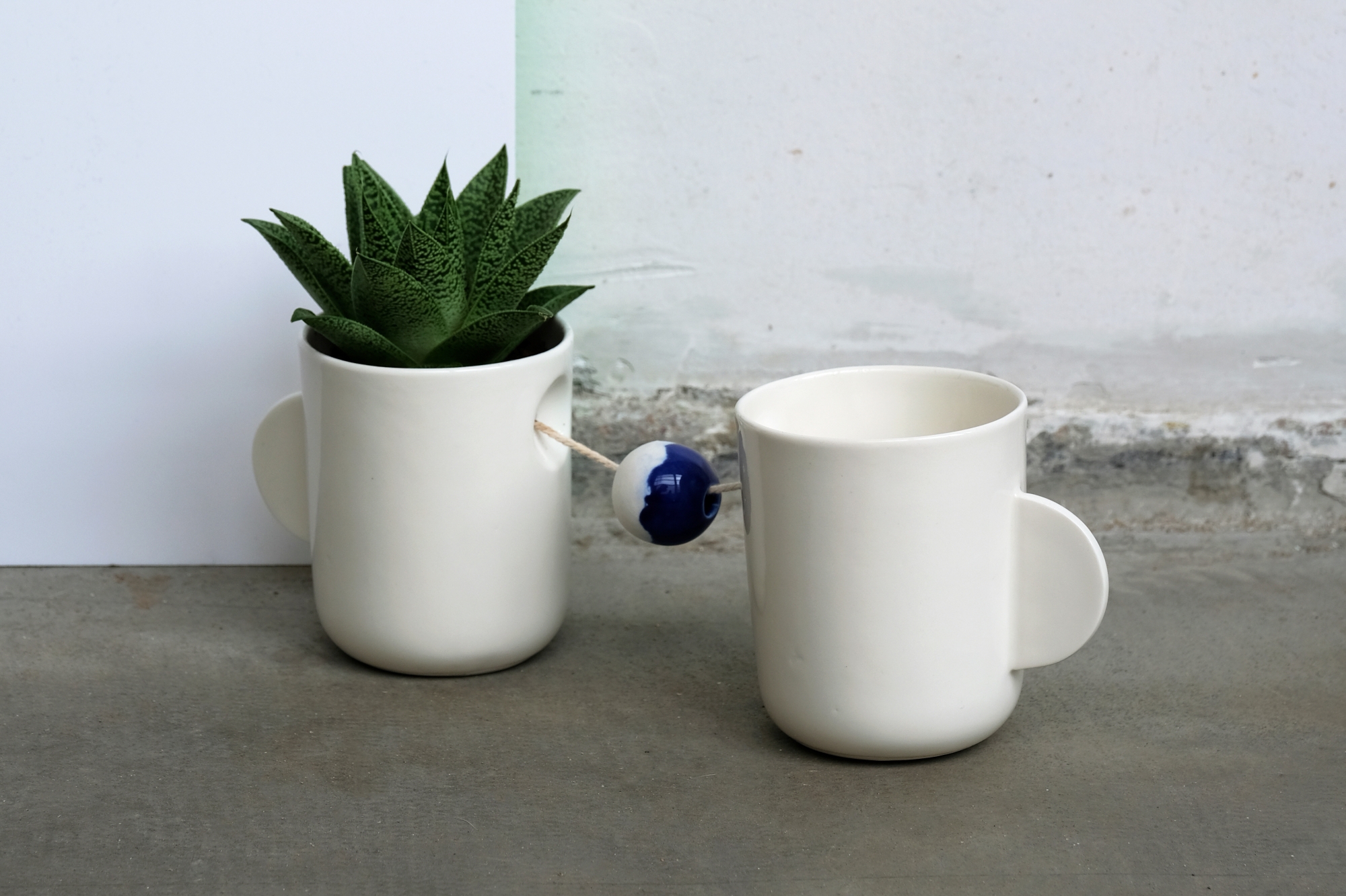 Dwa kubki ceramiczne zaprojektowane przez Katarzynę Sadowską; kubki są połączone ze sobą sznurkiem, w jednym z nich znajduje się roślinka