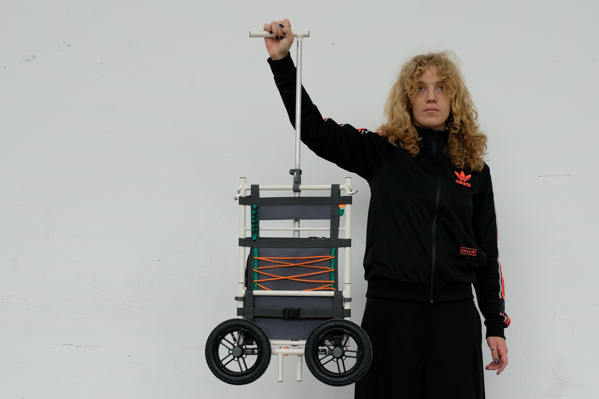 A woman holding a folded transport trolley in one hand by its extended T-handle. The trolley features a white frame, dark grey fabric panels laced with an orange bungee cord, and two large black wheels.