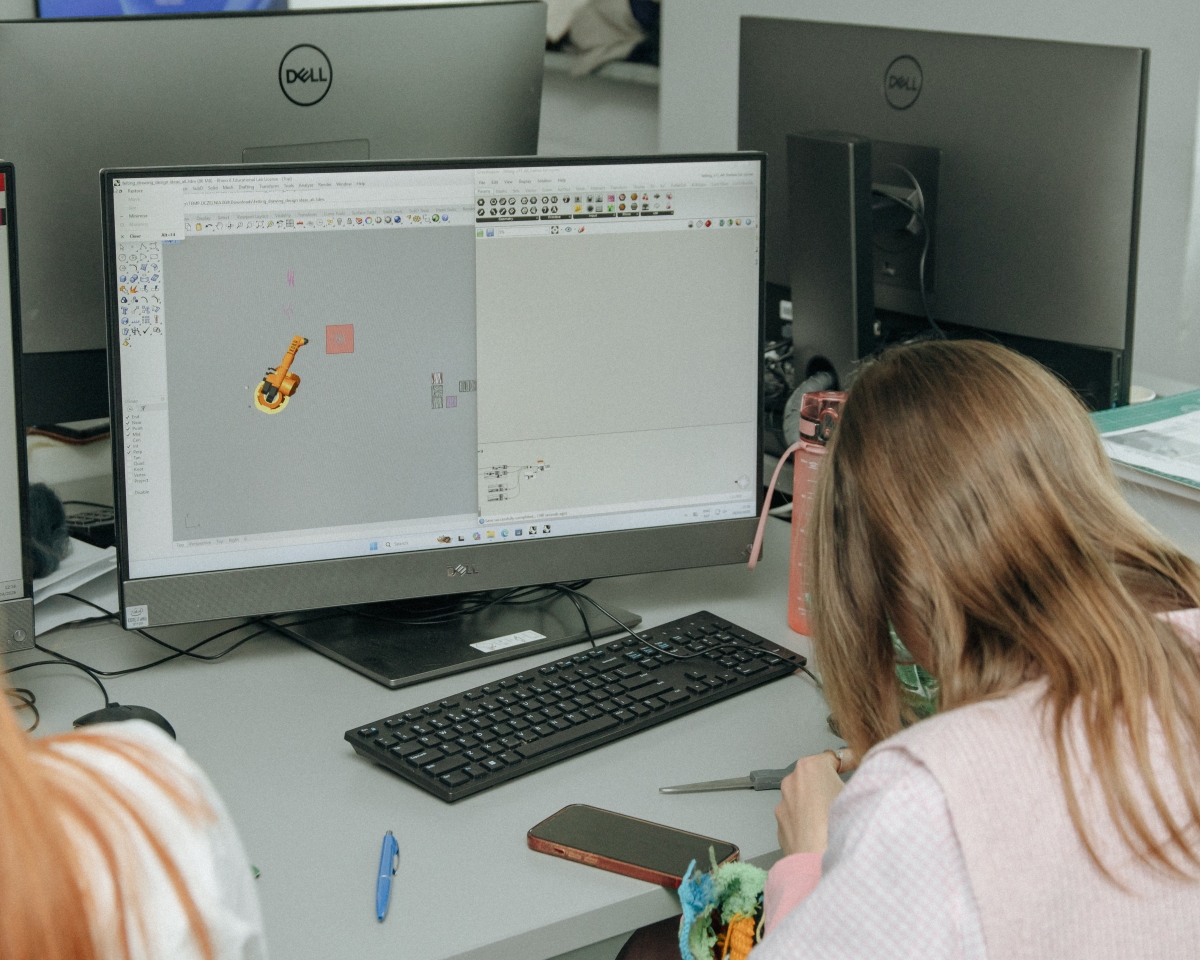 A young woman sitting at a computer. The screen displays the programming interface for an industrial robot arm.