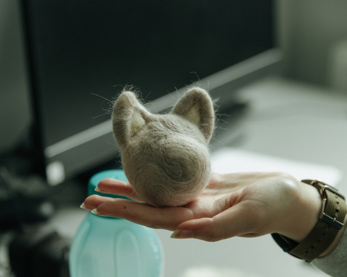 A hand holds a small, three-dimensional object made of light-colored felt that resembles an animal's head with protruding ears.