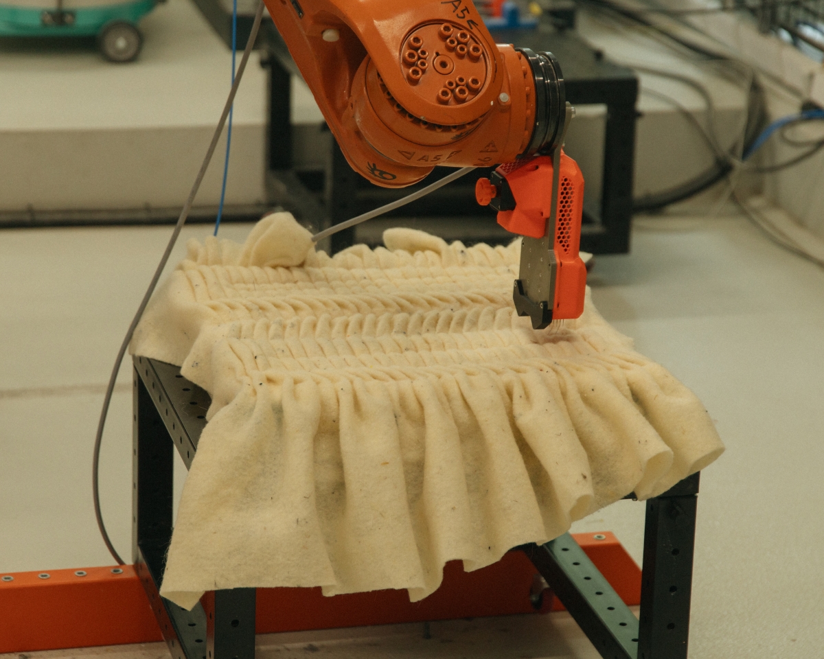 A robot arm equipped with a needle head working on a large piece of light-colored felt with a dense, regularly gathered, and pleated texture.