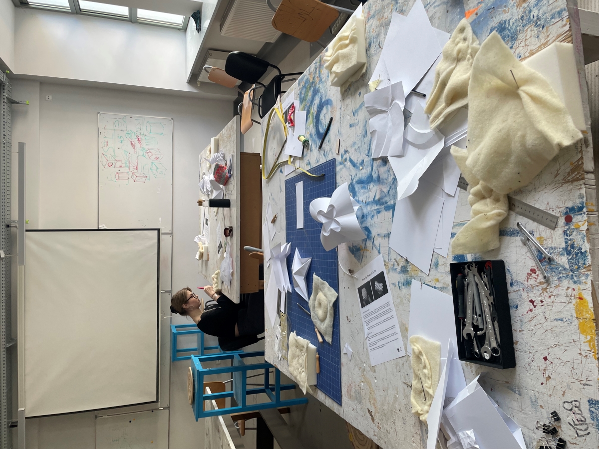 In the foreground, a workbench with pieces of felt and paper. In the background, a workshop participant sits at another table.
