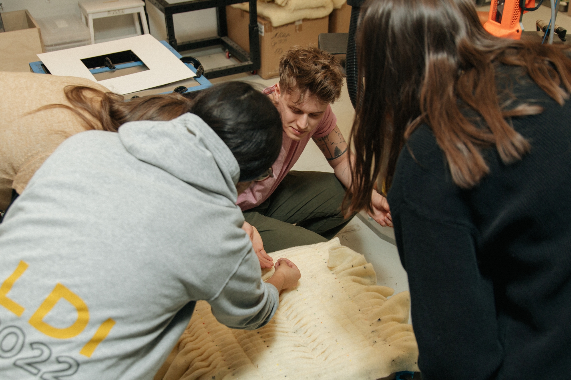 Three workshop participants are leaning over a large, pleated sheet of light-colored felt. In the background, a part of a KUKA robot arm and other workshop equipment are visible.