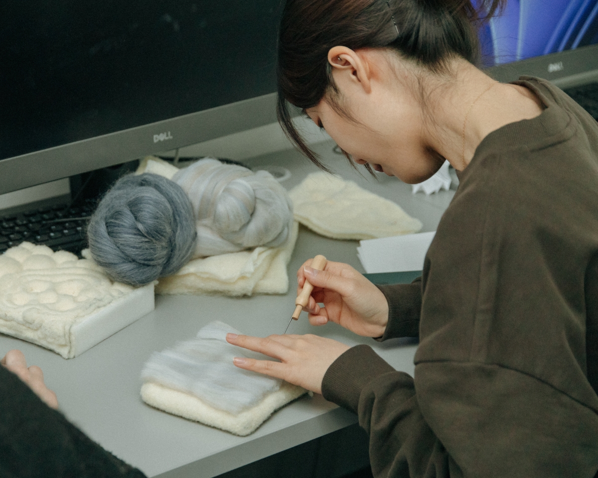  workshop participant felting light-colored wool using a tool. Next to it on the desk are rolled skeins of darker roving wool.