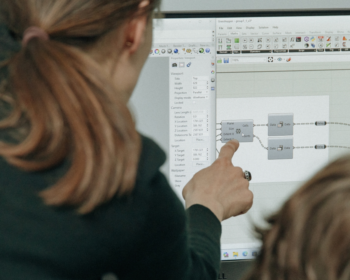 A close-up of a computer monitor showing the interface of an algorithmic design program. A person's hand is pointing a finger at a specific block with parameters.