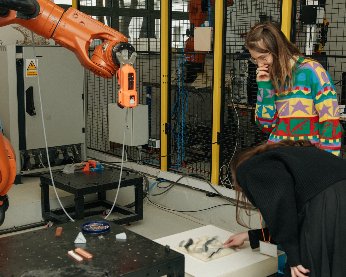 Two workshop participants leaning over a felt sample lying on a tabletop. Right next to it is a large industrial robot arm with a tool mounted at the end.