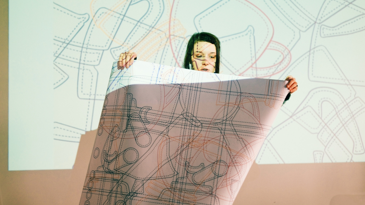 A School of Form student stands in front of a screen during a presentation, holding a sheet of paper with a printout of her project. Both the slide and the sheet of paper feature an illustration depicting a pattern of overlapping outlines of cups and coffee pots.