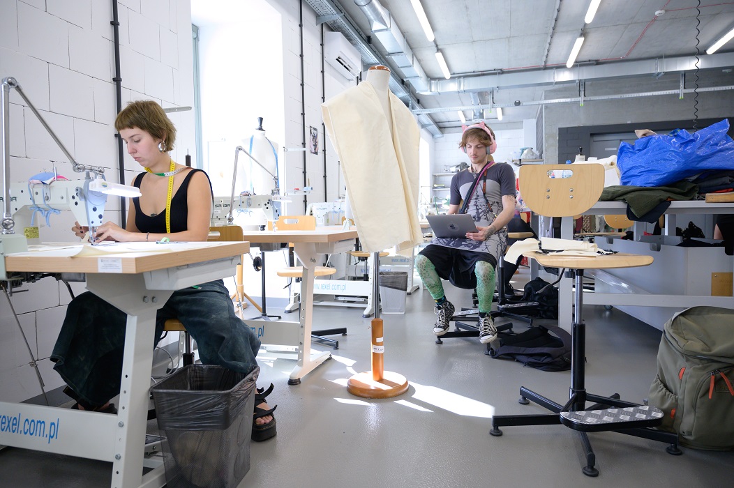 A student at a sewing machine in a sewing studio. In the distance, a student works on a laptop.